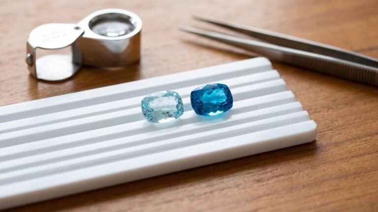 Aquamarine and blue topaz gemstones side by side on a white grooved sorting tray beside a loupe and tweezers