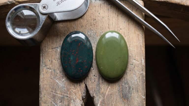 Polished bloodstone with red inclusions next to a plain green jasper cabochon on a wooden jeweler bench