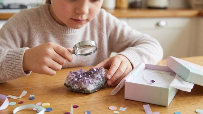 Young child examining an amethyst geode with a magnifying glass beside a delicate birthstone bracelet in a gift box