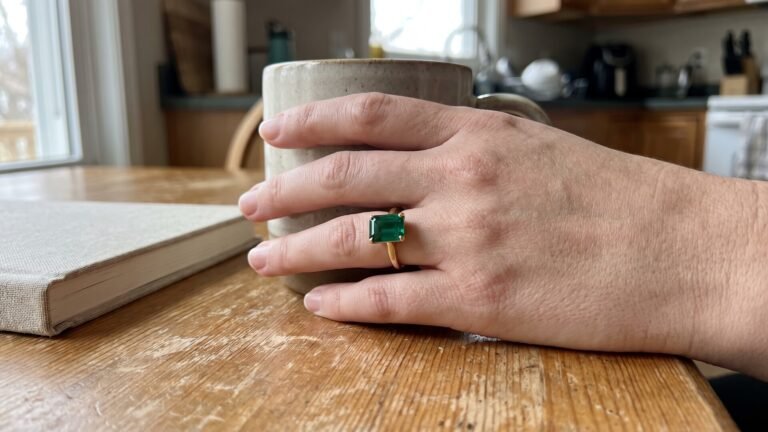 Woman wearing a gold emerald engagement ring while holding a ceramic mug at a kitchen table