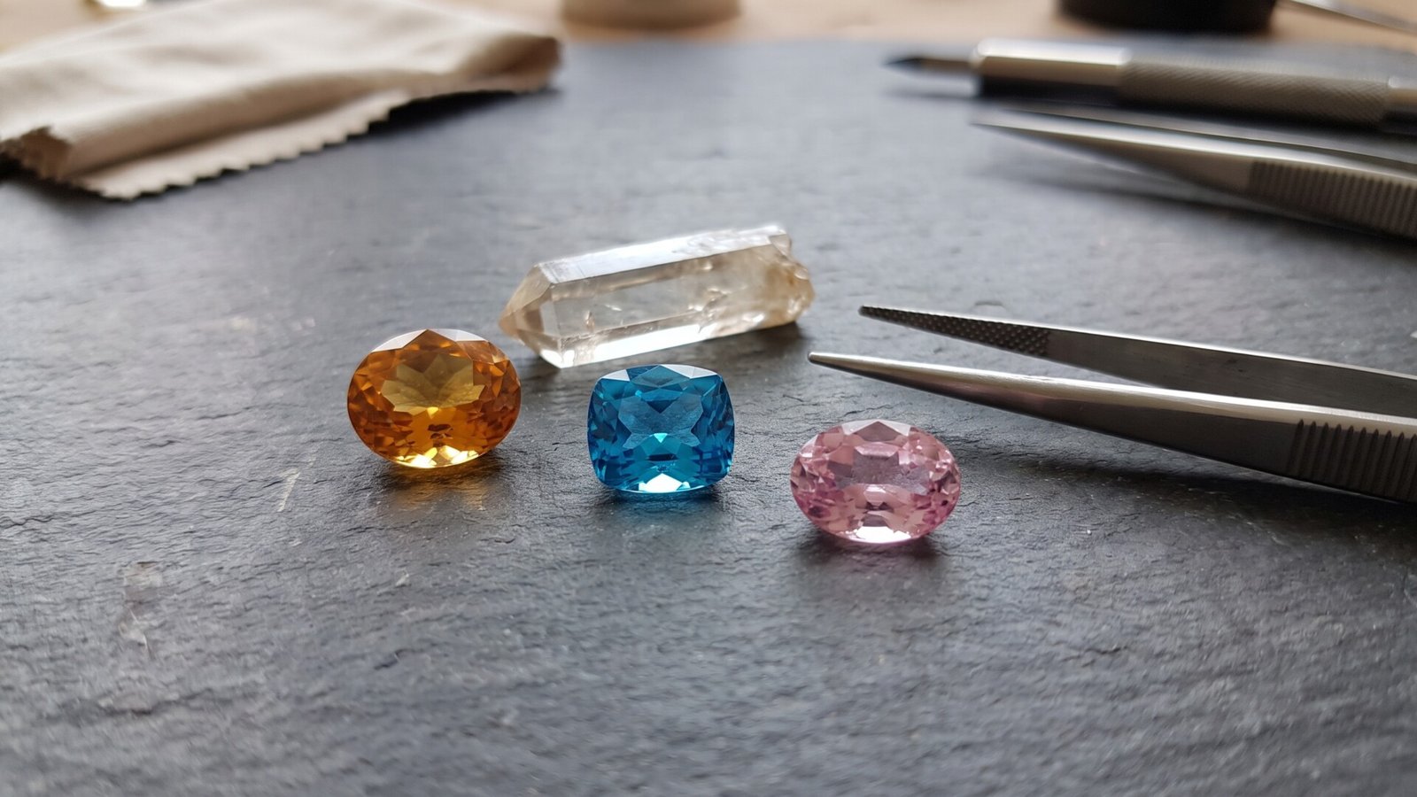 Faceted yellow, blue, and pink topaz gemstones displayed with a raw crystal point and jeweler tweezers