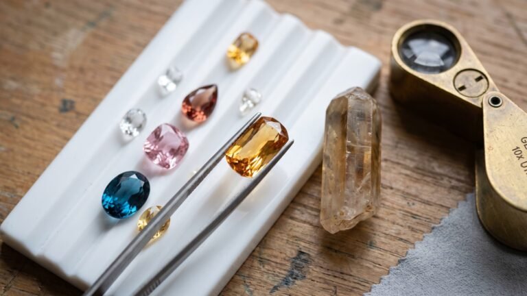 Faceted topaz gemstones on a white grading tray beside tweezers holding an imperial topaz and a raw crystal