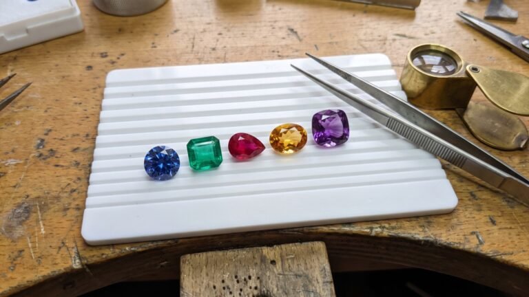 Five loose faceted gemstones in different cuts and colors arranged in a row on a white sorting tray