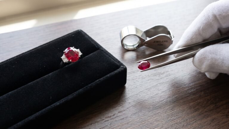 Jeweler holding a loose faceted ruby with tweezers next to an oval ruby ring and loupe on a wooden surface