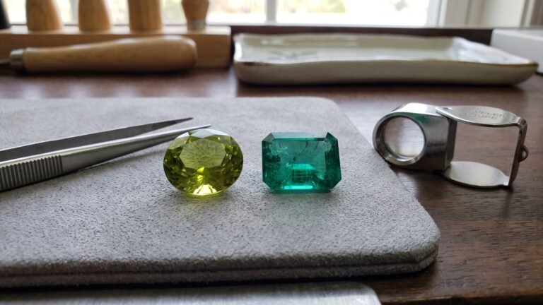 Faceted oval peridot and emerald-cut emerald on a jeweler's grading pad beside tweezers and a loupe
