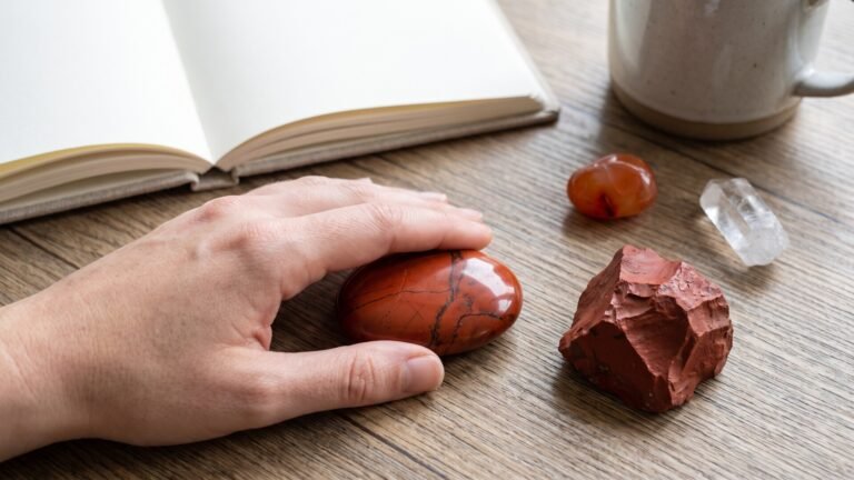 Hand resting on a polished red jasper stone beside a raw crystal chunk and open journal on a wooden desk