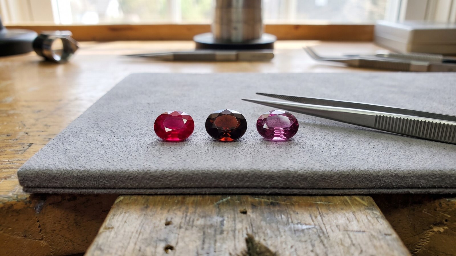 Three faceted oval gems—a bright red ruby, dark garnet, and pinkish tourmaline—arranged on a jeweler's pad
