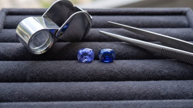 Faceted oval tanzanite and blue sapphire side by side next to a jeweler loupe and tweezers